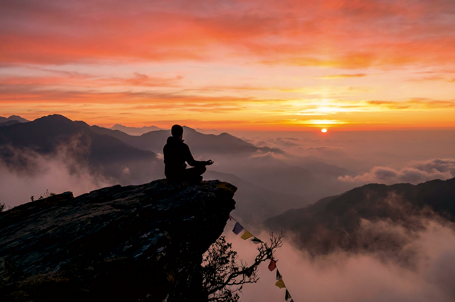 Person meditating in lotus position at sunrise overlooking misty mountains